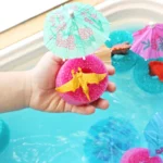 Child's hand holds a bright pink sponge with a small yellow dragon on it, with colorful paper umbrellas in a turquoise bath scene in the background.
