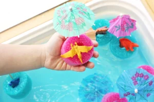Child's hand holds a bright pink sponge with a small yellow dragon on it, with colorful paper umbrellas in a turquoise bath scene in the background.
