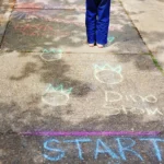 Child stands barefoot on a sunlit sidewalk, seen from behind, wearing a red shirt and blue pants amid colorful chalk drawings and writing on the ground.