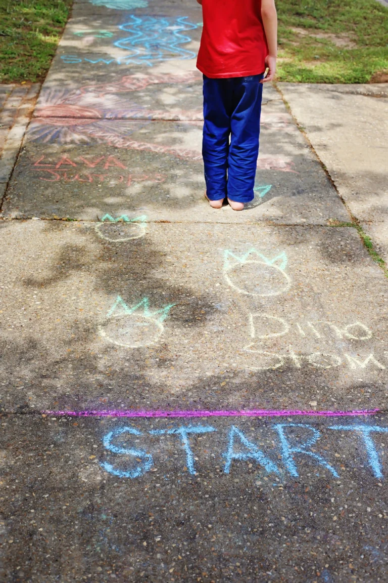 Child stands barefoot on a sunlit sidewalk, seen from behind, wearing a red shirt and blue pants amid colorful chalk drawings and writing on the ground.
