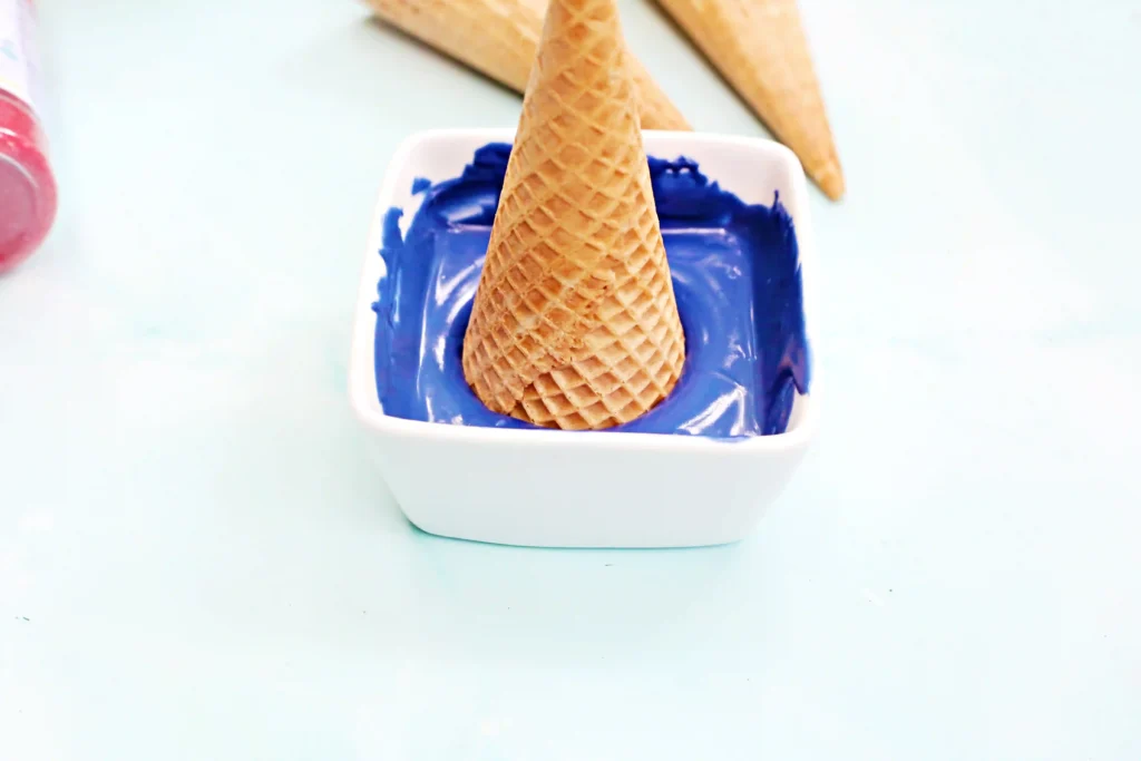 Close-up of a waffle cone standing upright in a white cup with melted blue candy melts around it.