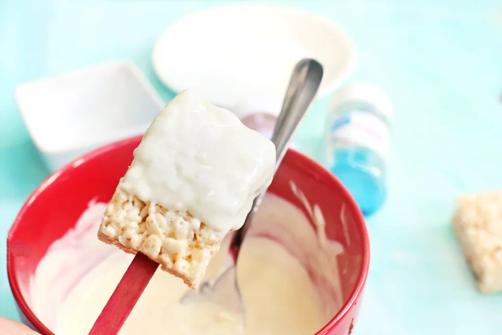 Patriotic Rice Krispie Pops - Cultivating Brilliant Minds Close-up of a rice krispie treat with melted white chocolate on the top half being held over a red bowl with a spoon nearby, turquoise background behind.