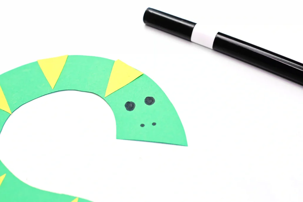 Close-up of a green curved paper strip with yellow triangles and black dots, next to a black marker on a white background for this s is for snake paper craft