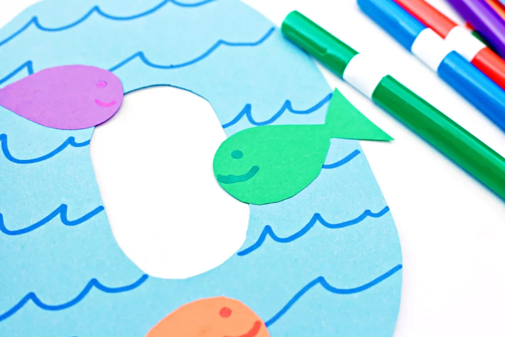 a letter O cut out of blue paper with blue waves drawn on and colorful paper fish glued on top