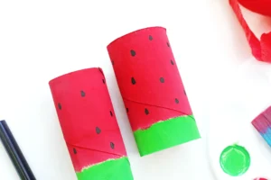 Watermelon-inspired craft tubes: red toilet paper rolls with black seeds and green bottoms on a white surface.