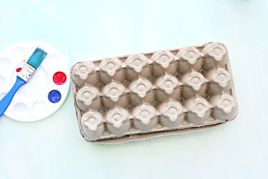 Blue paintbrush resting on a white palette with red and blue paint, beside a brown egg carton on a pale blue surface.
