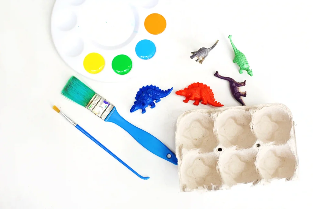 paint brushes next to a paint palette with orange, blue, green, and yellow paint, toy dinosaurs, and a cardboard egg carton for the dinosaur egg carton craft