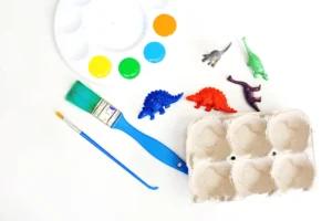 paint brushes next to a paint palette with orange, blue, green, and yellow paint, toy dinosaurs, and a cardboard egg carton