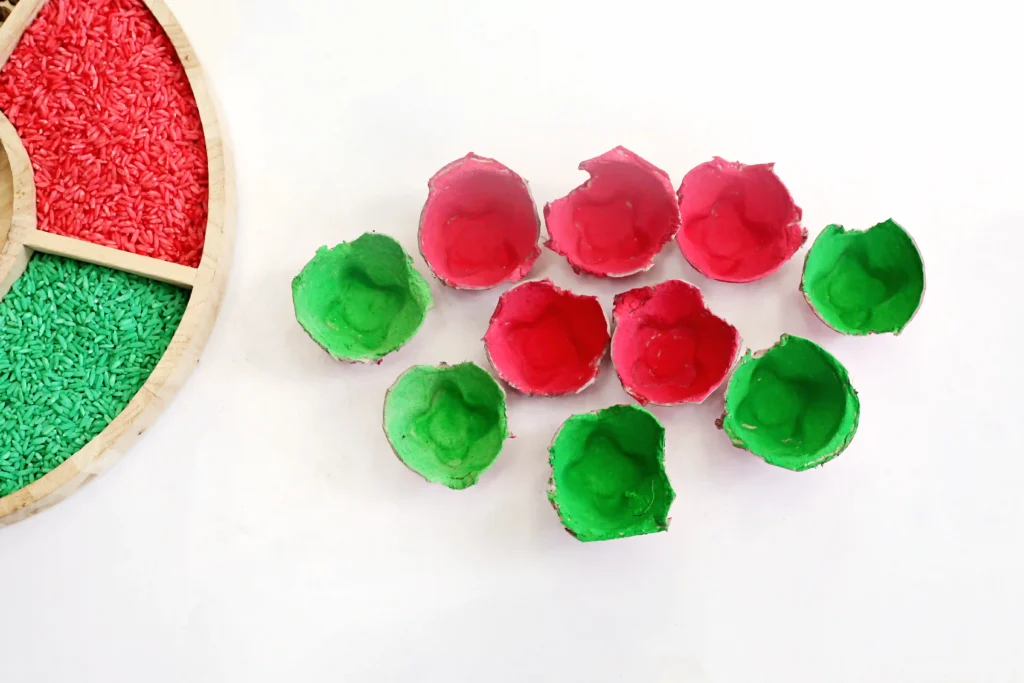 Egg Carton Watermelon Fine Motor Activity - Cultivating Brilliant Minds Colorful craft supplies: red and green dyed rice in a wooden tray beside dyed egg carton cups on a white background.