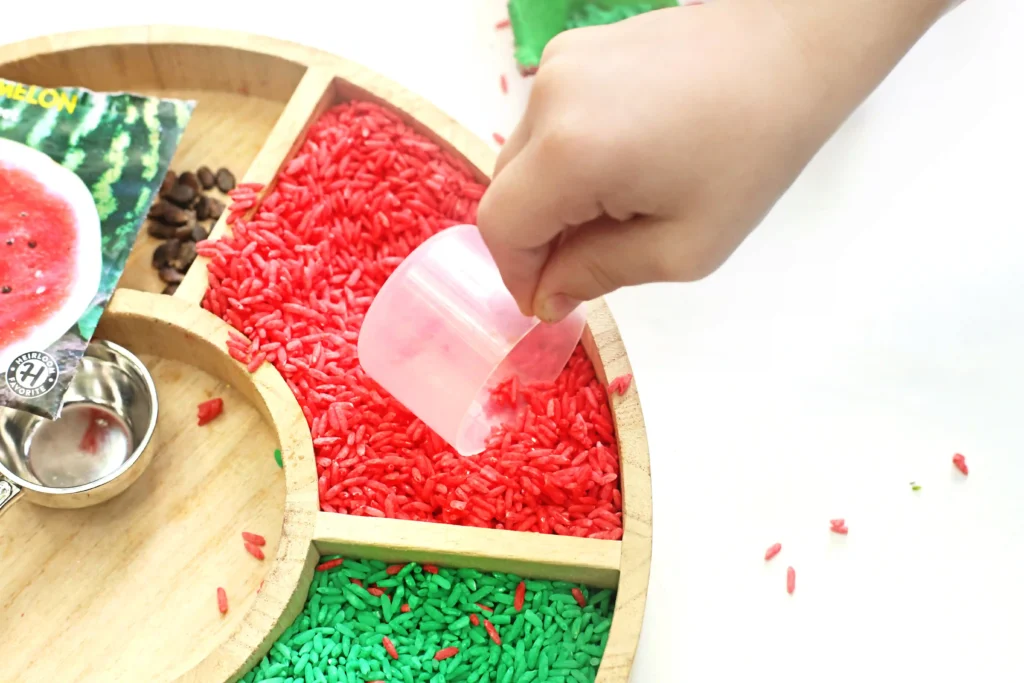 Egg Carton Watermelon Fine Motor Activity - Cultivating Brilliant Minds Close-up of a wooden tray with red and green colored rice grains; a hand scoops up red rice