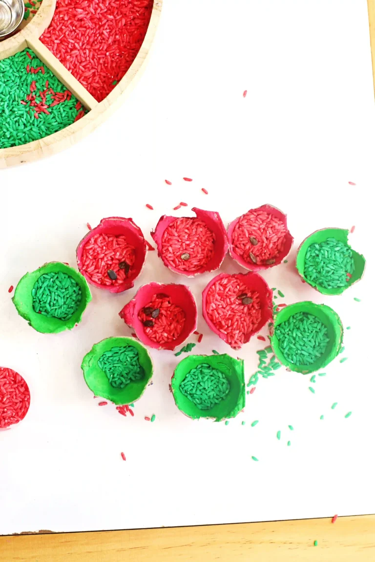 Colorful red and green rice grains in painted egg carton cups arranged on a white surface, with a wooden tray in the corner and scattered pieces around.