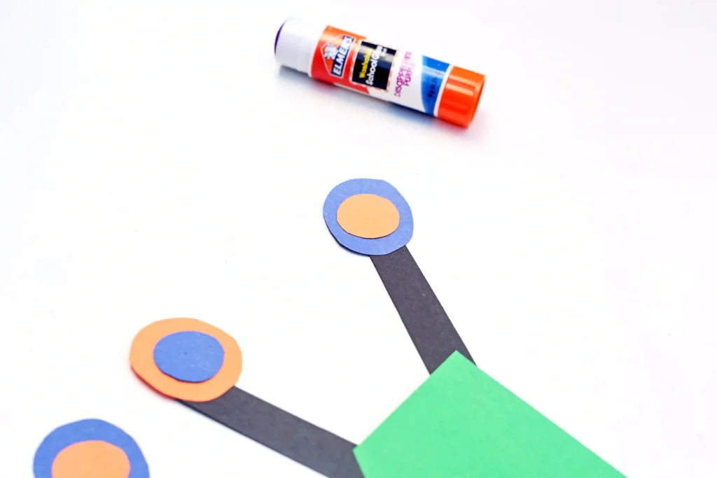 Glue stick beside orange-on-blue paper circles arranged along a black strip on a white background.
