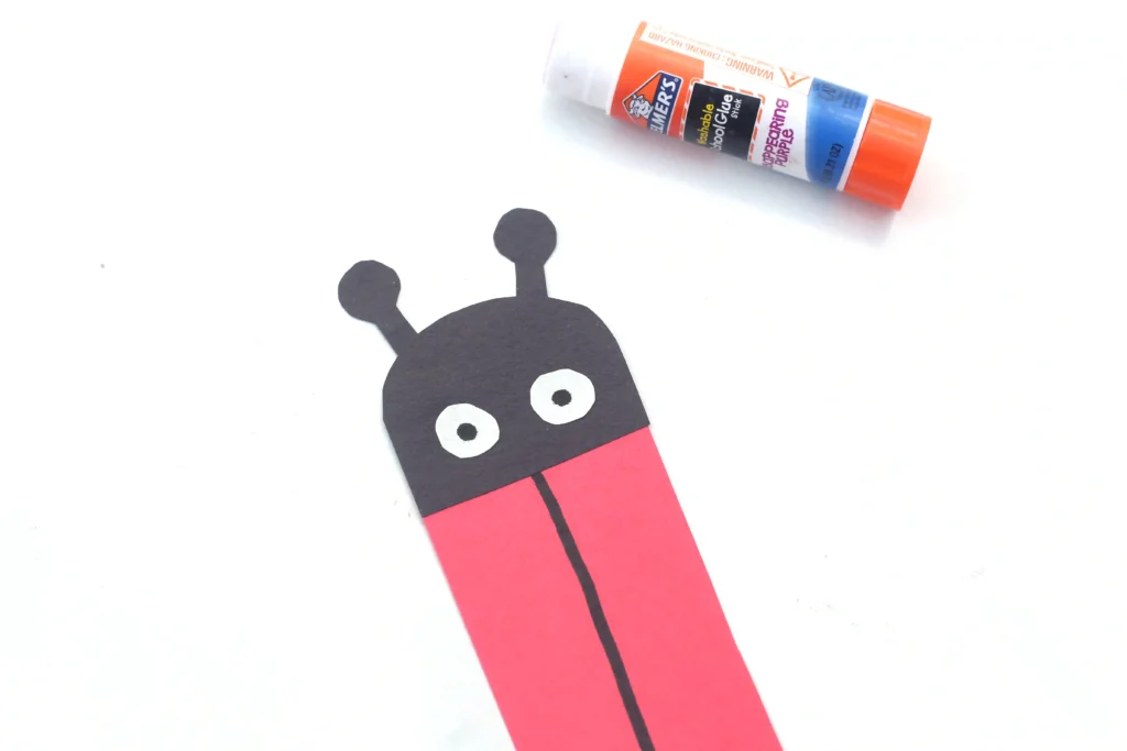 Paper cut-out of a ladybug with red body shaped like an L and two antennae on a white background, beside a glue stick for craft projects.