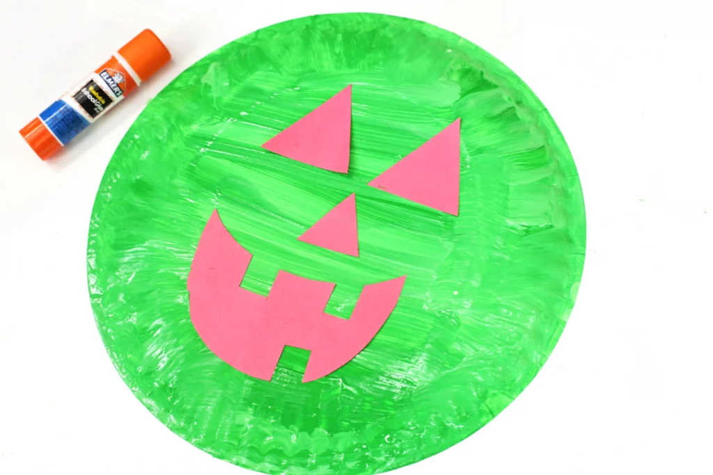 Green circular paper plate painted with a pink jack-o'-lantern face made of triangular cutouts and a glue stick nearby