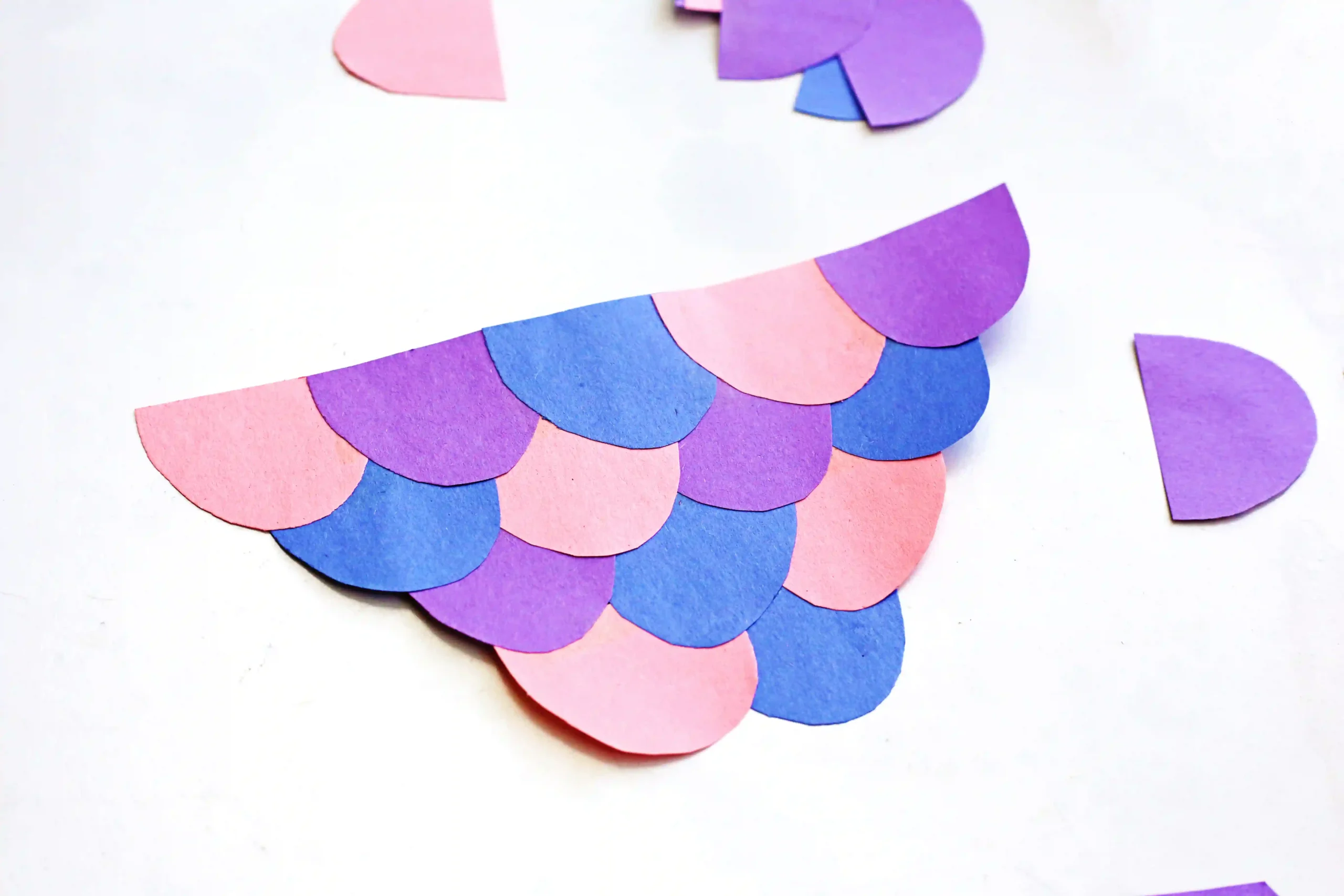 Colorful paper cut-out scales in pink, purple, and blue forming a fish tail on a white background.