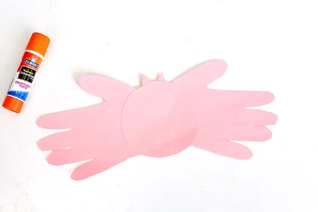 pink construction paper hand cut outs glued onto a circle laid on a white surface with an orange Elmer's glue stick in the corner, suggesting a craft project.