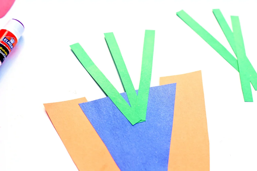 Colorful paper craft with orange and blue triangles and green paper strips arranged on a white background; glue bottle appears on the left.