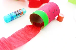 Colorful paper craft resembling a watermelon wedge with pink body, green rind, black seeds, red tissue, and a nearby glue stick.