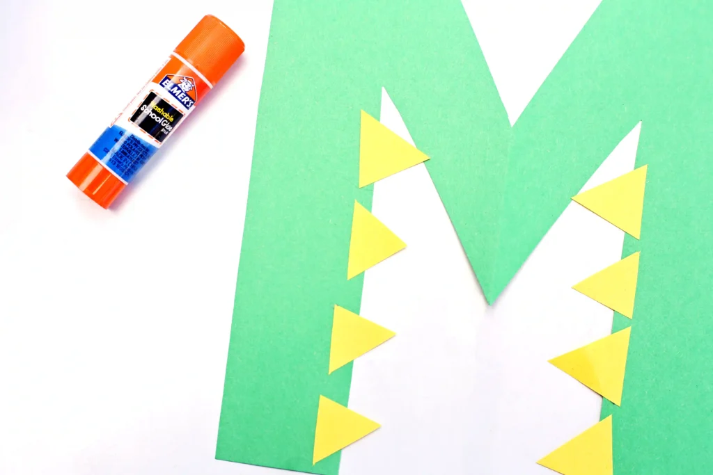 Orange glue stick on white surface with a green letter M and yellow triangles glued on