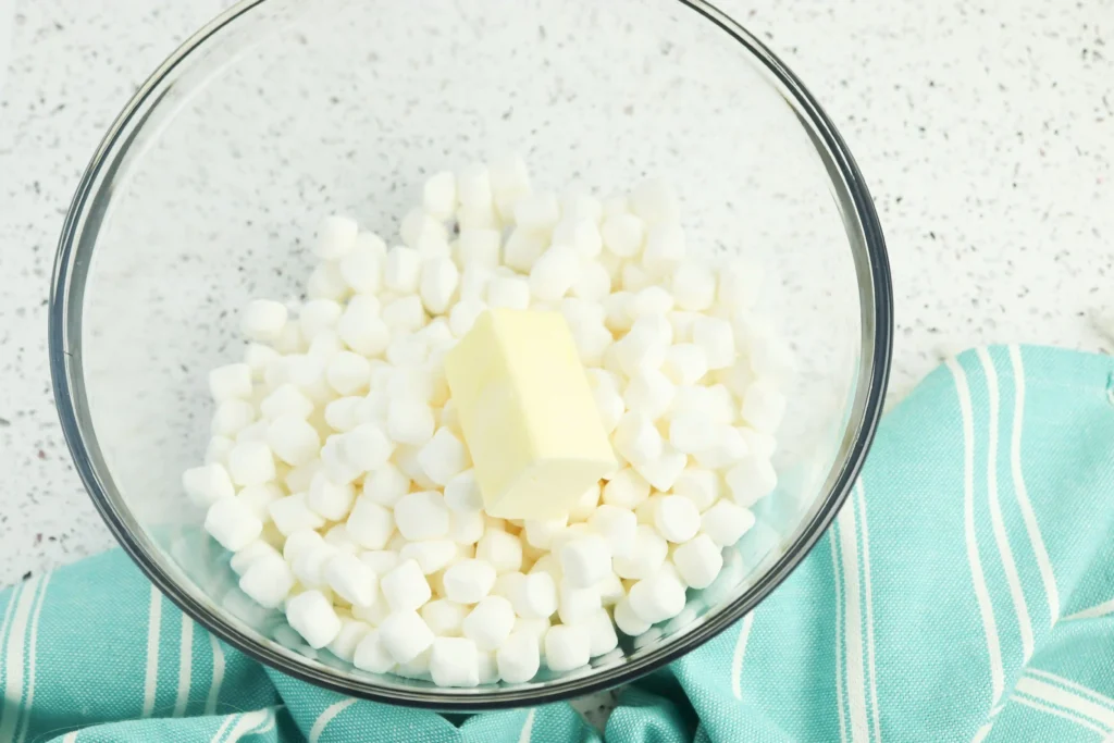 Glass mixing bowl filled with white mini marshmallows and butter cubes, set on a speckled countertop with a turquoise striped cloth nearby