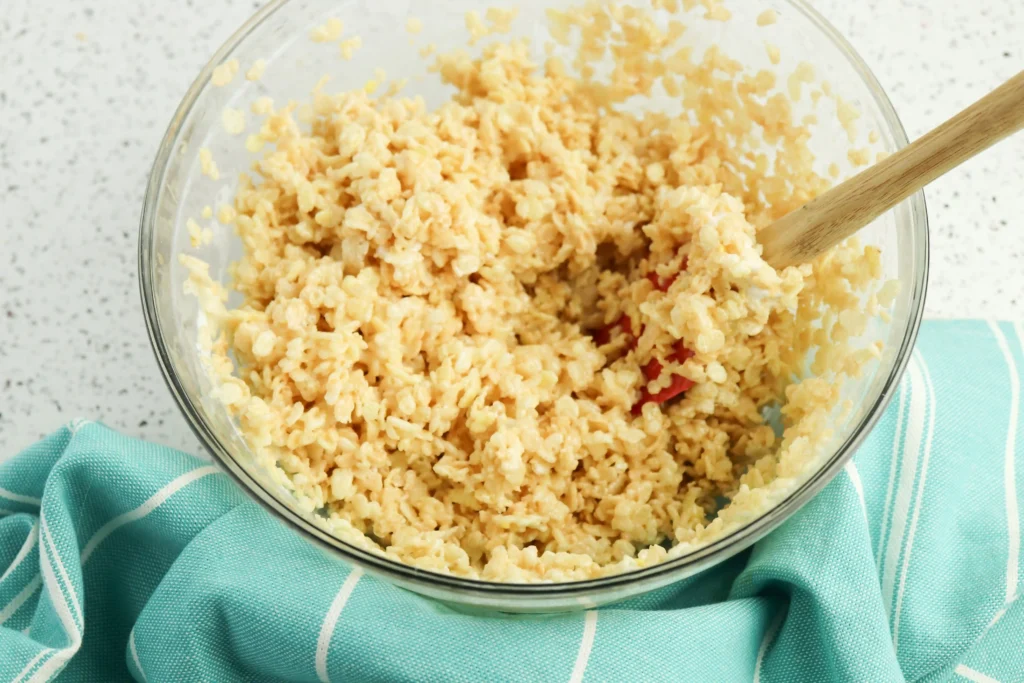 rice krispie batter in a glass bowl with a wooden spoon, resting on a turquoise striped cloth