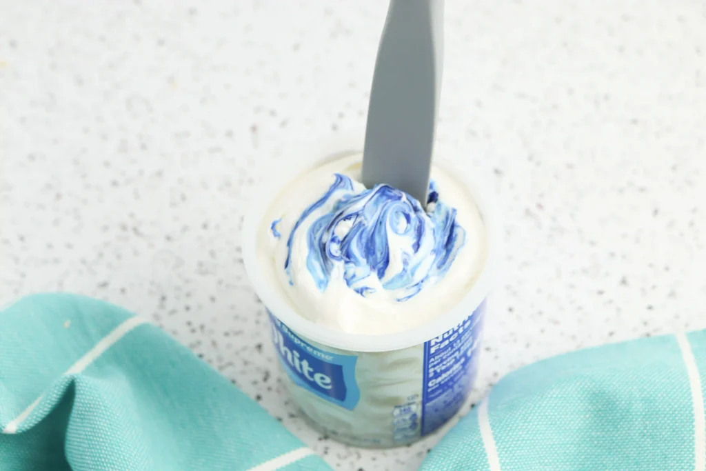 Cup of vanilla icing with blue swirl food coloring and a grey spoon piercing the center on a speckled countertop.