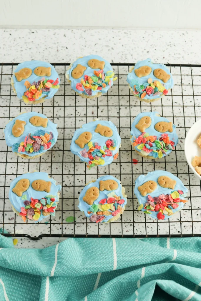 Blue-frosted cupcakes with goldfish crackers  and fruity pebble cereal toppings on a cooling rack.