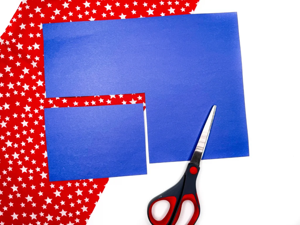Blue scissors and blue craft paper pieces on a red star-patterned background (crafting setup).
