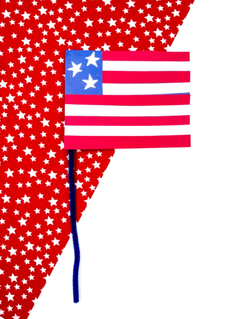an american flag craft made out of construction paper with a pipe cleaner pole on a red and white star background