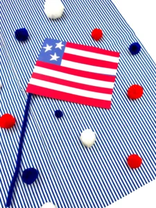 an american flag craft made out of construction paper with a pipe cleaner pole on a blue and white striped background with red, white, and blue pom poms scattered around