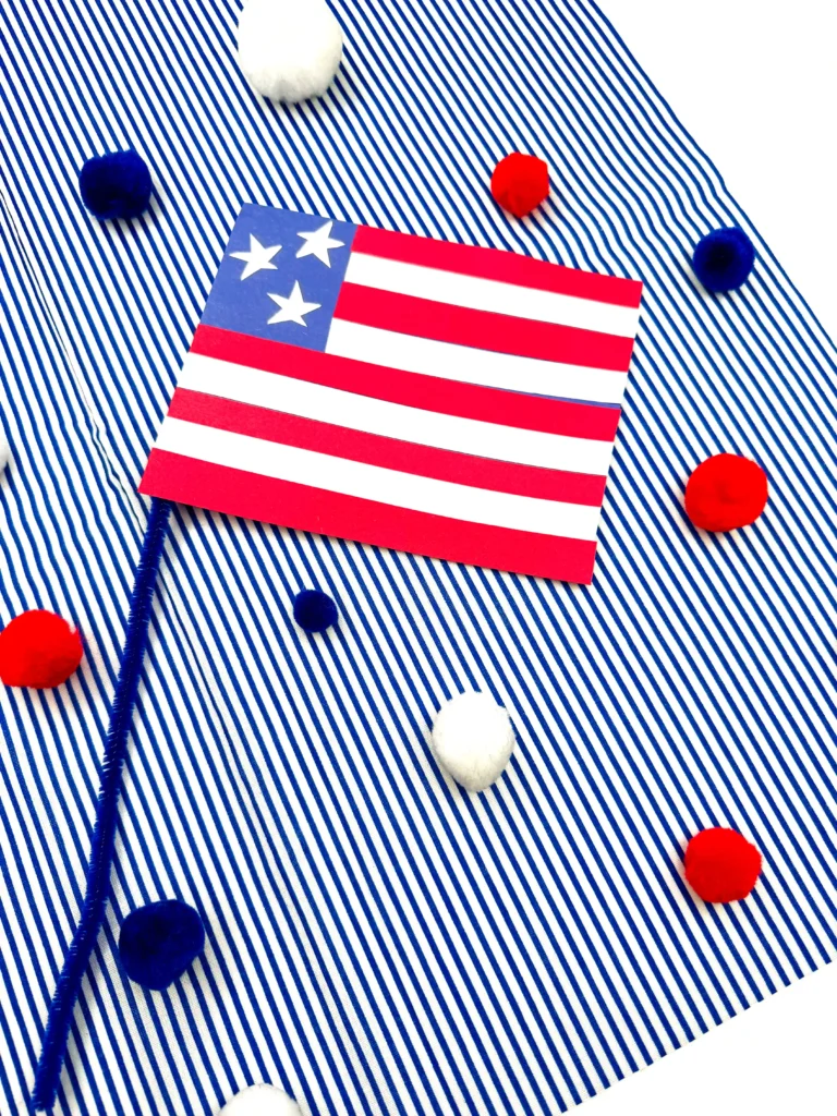 an american flag craft made out of construction paper with a pipe cleaner pole on a blue and white striped background with red, white, and blue pom poms scattered around