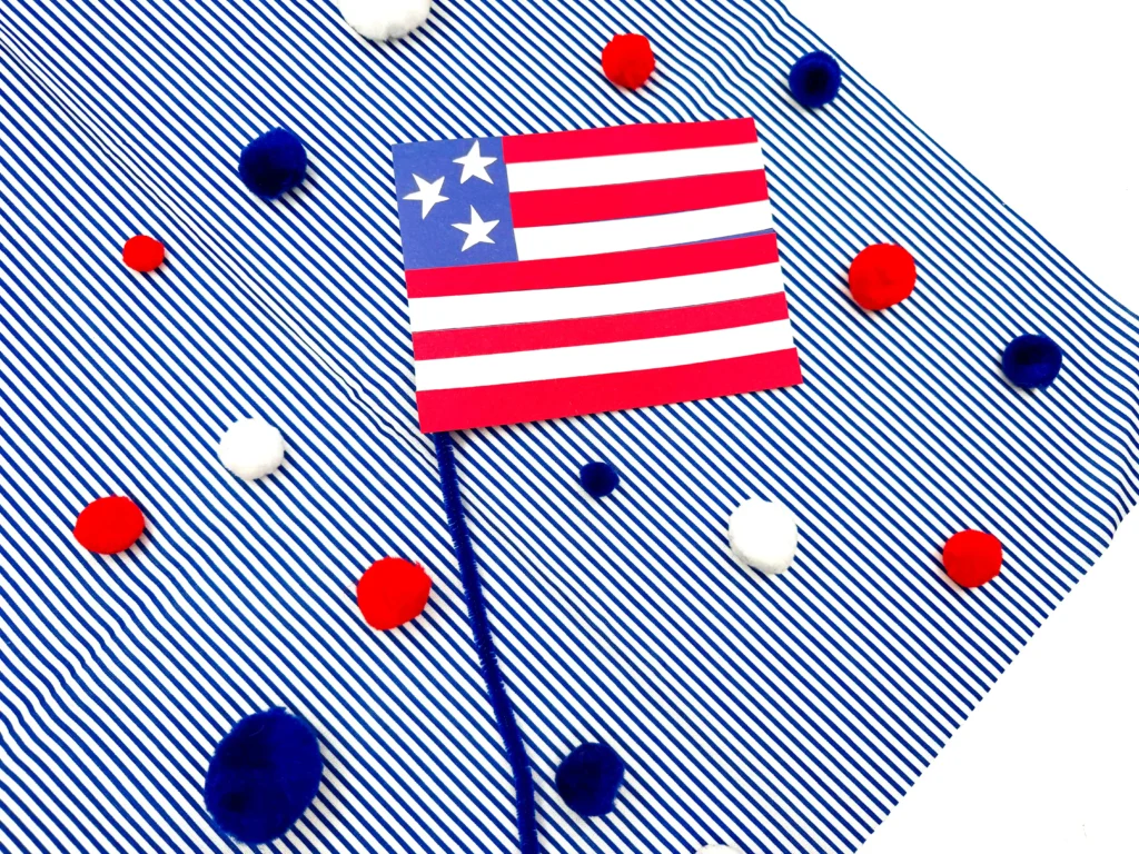 an american flag craft made out of construction paper with a pipe cleaner pole on a blue and white striped background with red, white, and blue pom poms scattered around