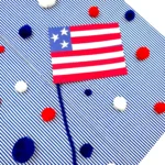 an american flag craft made out of construction paper with a pipe cleaner pole on a blue and white striped background with red, white, and blue pom poms scattered around