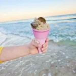 Child's hand in a yellow sleeve holds a pink ice cream cone with sand and seashells inside at the beach, with calm ocean and sunset in the background.