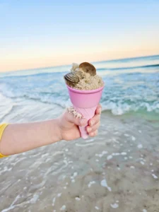 Child's hand in a yellow sleeve holds a pink ice cream cone with sand and seashells inside at the beach, with calm ocean and sunset in the background.