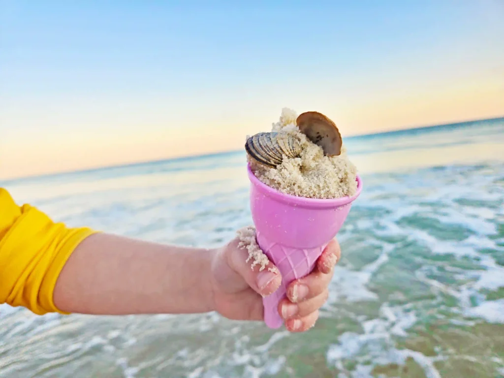 Child's hand in a yellow sleeve holds a pink ice cream cone with sand and seashells inside at the beach, with calm ocean and sunset in the background.