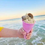 Child's hand in a yellow sleeve holds a pink ice cream cone with sand and seashells inside at the beach, with calm ocean and sunset in the background.