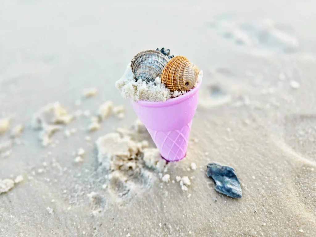 Pink plastic waffle cone with two seashells on top, set against a sandy beach.