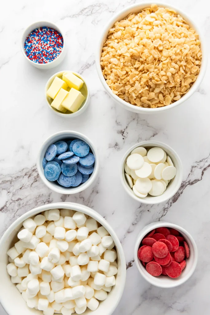 ingredients for the patriotic rice krispie treats - Top-down view of bowls with baking toppings: cornflakes, butter cubes, red/white/blue sprinkles, blue candies, white chocolate chips, mini marshmallows, and red candy discs.