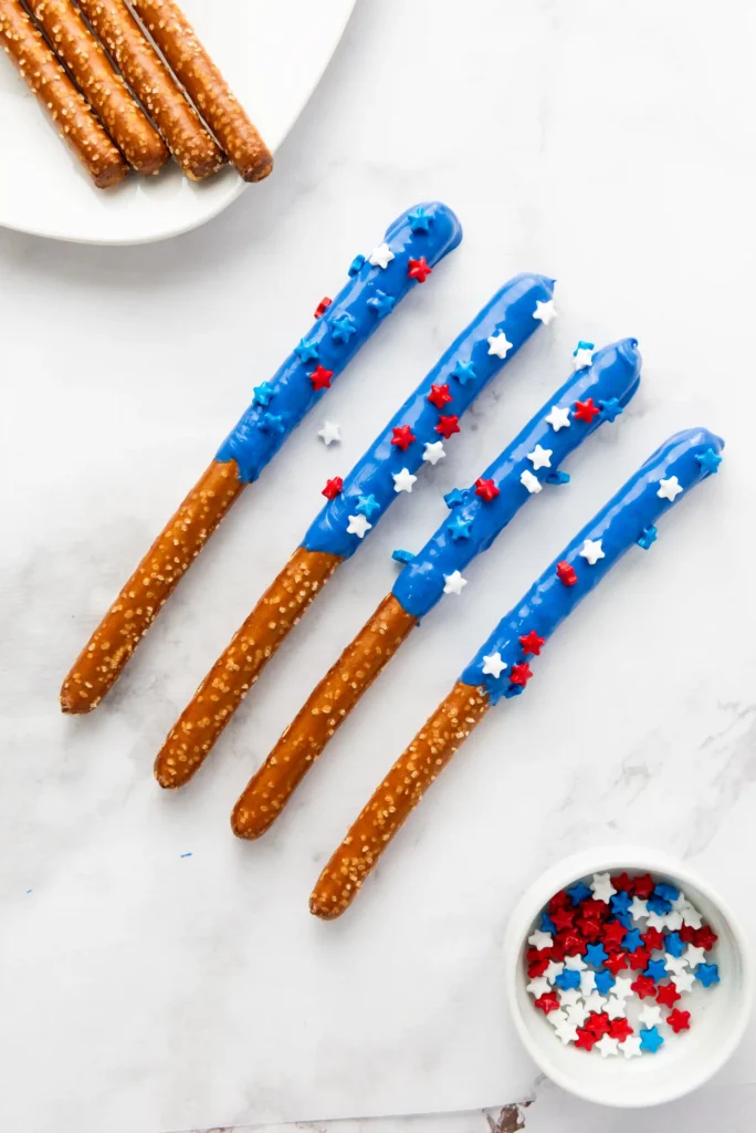 Blue chocolate-dipped pretzel rods with red, white, and blue star sprinkles on a white surface; extra pretzels on a plate nearby and a bowl of sprinkles in the corner.