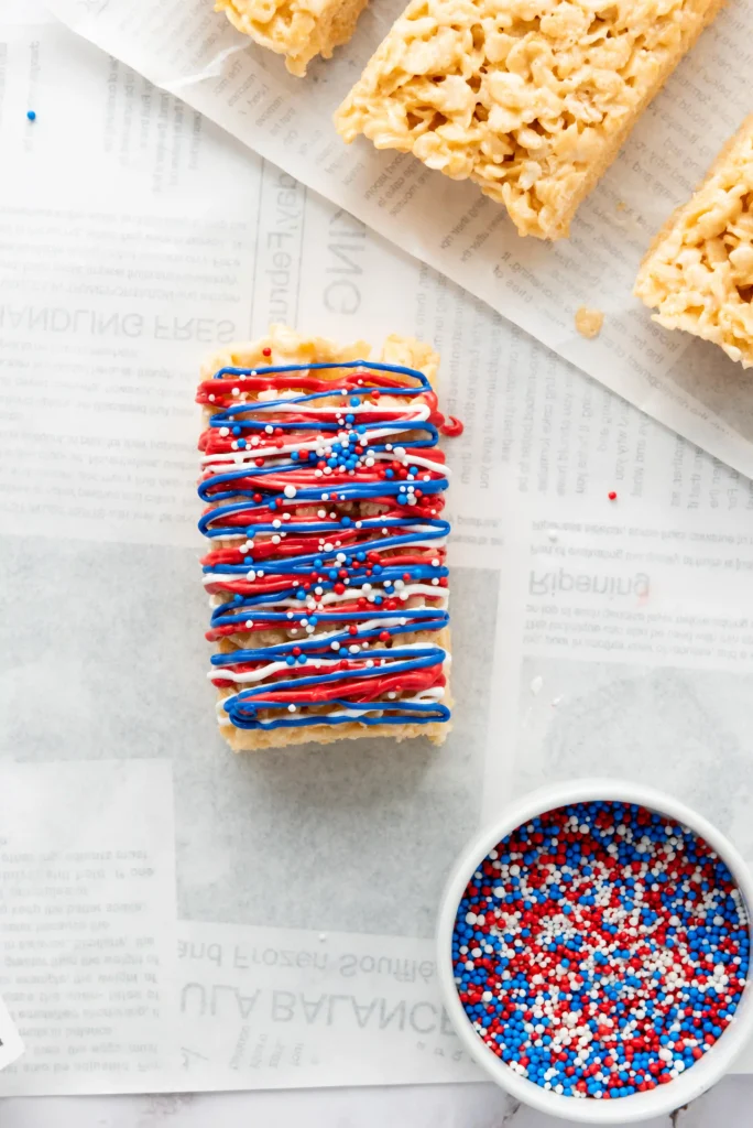 Red, white, and blue drizzled dessert bar with sprinkles on parchment paper and a bowl of matching sprinkles nearby.