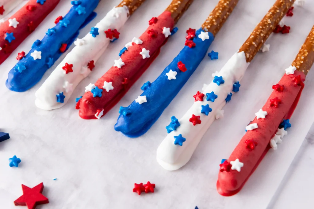Red, white, and blue pretzel rods coated in icing with white, blue, and red star sprinkles arranged on a marble surface (patriotic treats).