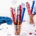 Patriotic snacks: red, white, and blue pretzel sticks with star sprinkles in glass jars on a white surface.