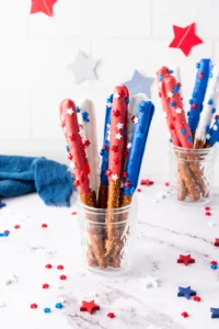 Patriotic snacks: red, white, and blue pretzel sticks with star sprinkles in glass jars on a white surface.