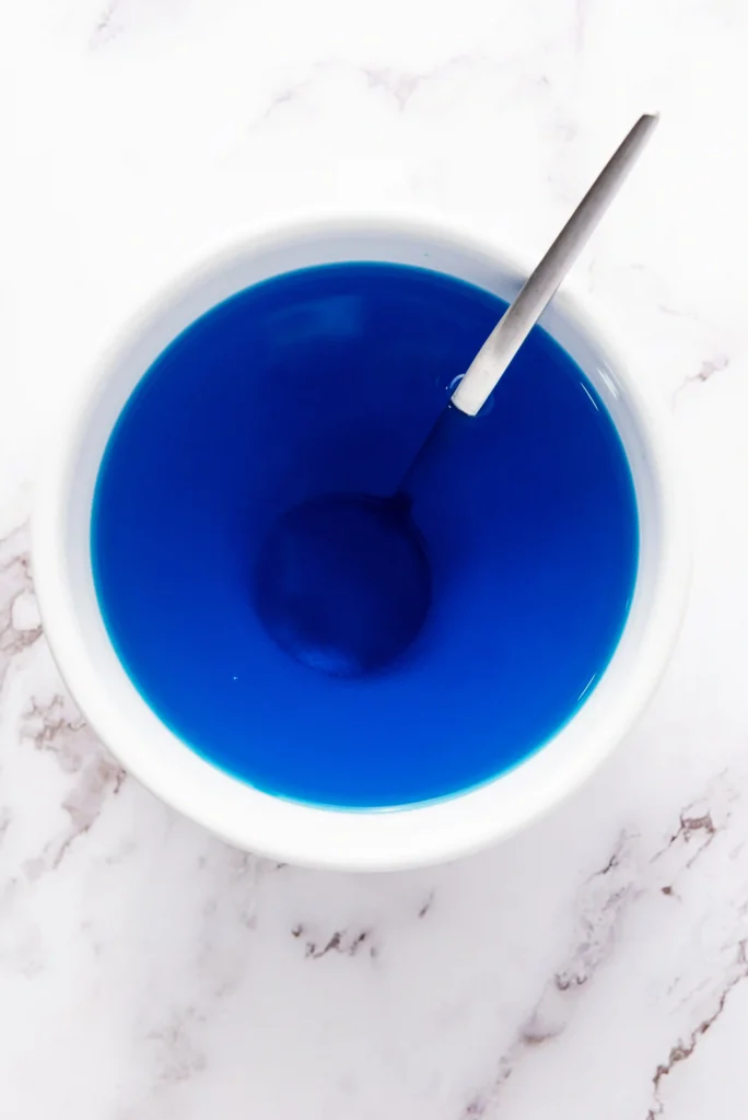 Top-down view of a white mixing bowl filled with bright blue jello liquid and a metal spoon resting inside.