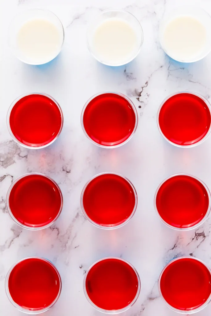 Overhead view of a grid of clear cups on a marble surface, with red jello in the bottom rows and white gelatin in the top row.