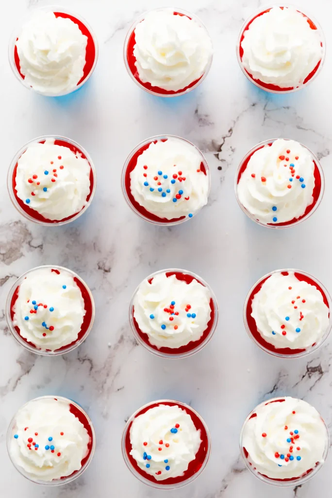 Top-down view of red jelly cups topped with whipped cream and blue and red sprinkles on a marble surface.