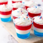 Patriotic layered dessert cups in red, white, and blue with whipped cream and red‑blue sprinkles on a marble surface.