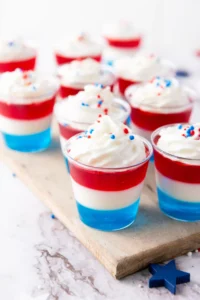 Patriotic layered dessert cups in red, white, and blue with whipped cream and red‑blue sprinkles on a marble surface.
