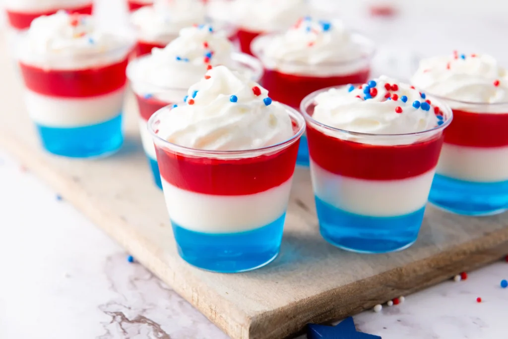 Patriotic desserts in a cup with red, white, and blue topped with whipped cream and sprinkles on a wooden board